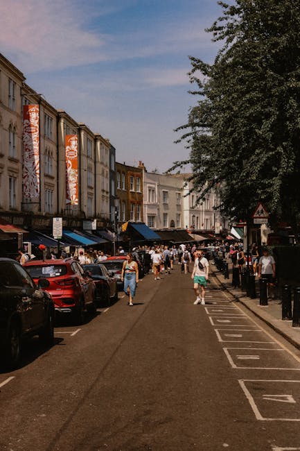 A busy street scene in Bermondsey with parked cars on the left side, including a red vehicle, and pedestrians walking along the sidewalk, some carrying shopping bags. On the right side, there is a row of white and brick buildings with large windows and signs, and a large leafy tree partially shading the pavement. The sky is partly cloudy with patches of blue. In the background, more people and storefronts are visible, indicating a vibrant shopping and social area. This scene captures the lively environment typical of Bermondsey street, where local removals and home relocation services, such as those provided by Man With a Van Bermondsey, are often necessary to facilitate furniture transport, packing, and moving logistics.