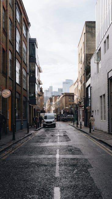 A narrow urban street scene on a cloudy day in Bermondsey with tall buildings lining both sides, including brick and modern white facades; the pavement is wet, indicating recent rain, and black bollards separate the sidewalk from the road. In the foreground, a white van is parked near the curb, with a few pedestrians walking along the street. The image captures the typical environment of Bermondsey, often involved in home relocation and furniture transport activities, relevant to services offered by Man With a Van Bermondsey, which specialises in removals and moving logistics within the area.