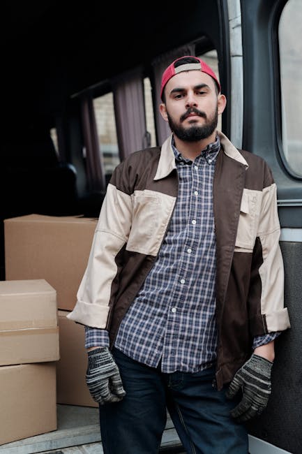 A man wearing a red cap, beige and brown jacket, and grey gloves stands inside a van used for home relocation, with cardboard boxes positioned behind him. The van's interior is visible with a dark ceiling and side panels, and the man appears to be part of a furniture transport or packing and moving service. Outside the open rear doors, a paved area is visible, indicating the loading process typical of house removals. The setting reflects professional belongings handling, aligning with house removal or moving services provided by Man With a Van Bermondsey, supporting efficient furniture transport and packing during a move.