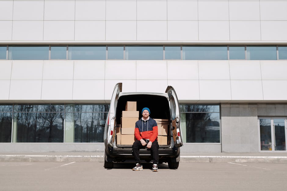 A man wearing a red and blue jacket, black pants, and a blue beanie stands with his arms crossed outside a silver cargo van parked on a paved area in front of a modern, white building with large windows and horizontal architectural lines. The back doors of the van are open, revealing an empty interior with cardboard boxes and packing materials, indicating a home relocation and furniture transport process. The van is positioned near a curb, and the sky above is clear with some light clouds. This scene depicts the loading or unloading phase of a professional moving service, consistent with house removal activities provided by Man With a Van Bermondsey, supporting orderly packing and moving operations.