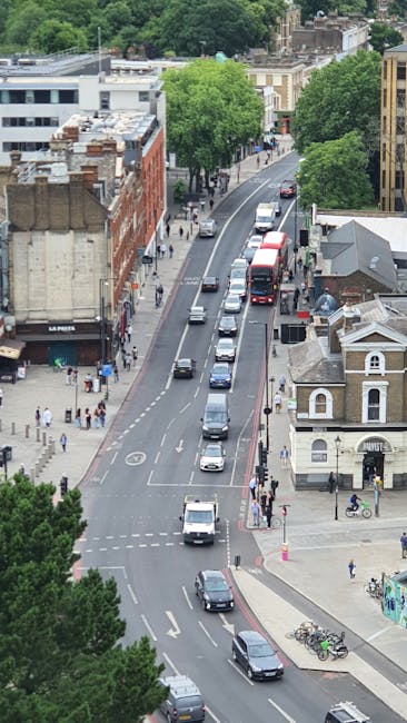 An aerial view of a busy urban street in Bermondsey with a mix of vehicles including cars, a red double-decker bus, and bicycles, alongside pedestrians walking on the sidewalks. To the right, there is a small white building with a grey roof, and further in the background, there are larger brick and modern buildings with rooftops and chimneys. Tall green trees line the street, providing shade and visual contrast to the buildings. The scene captures the typical city environment where house removals and furniture transport services by Man With a Van Bermondsey might operate, illustrating the busy street scene during daytime, with natural lighting highlighting movement and activity involved in home relocation and packing and moving processes.